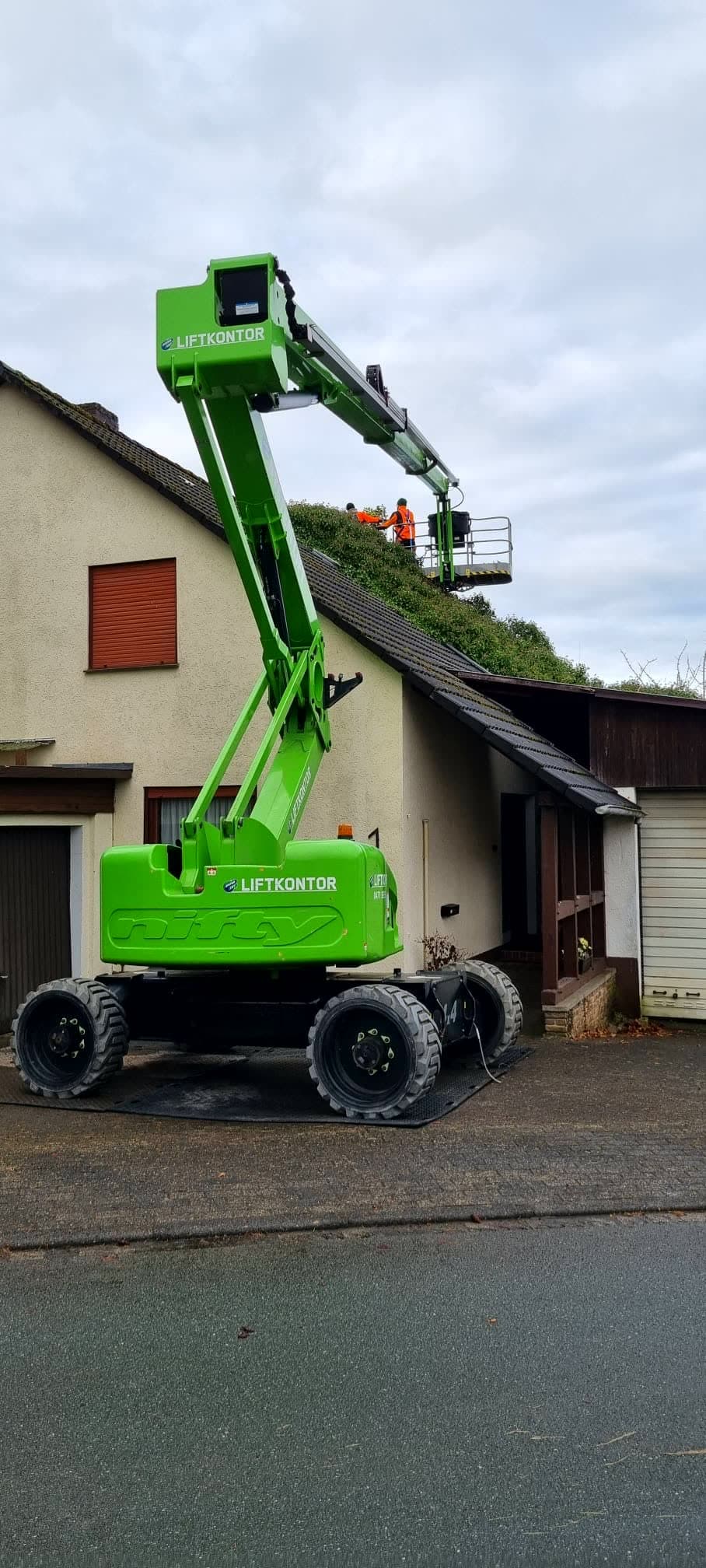 Grüne Teleskop-Arbeitsbühne vor einem Haus, oben im Korb arbeiten zwei Mitarbeiter am Dach