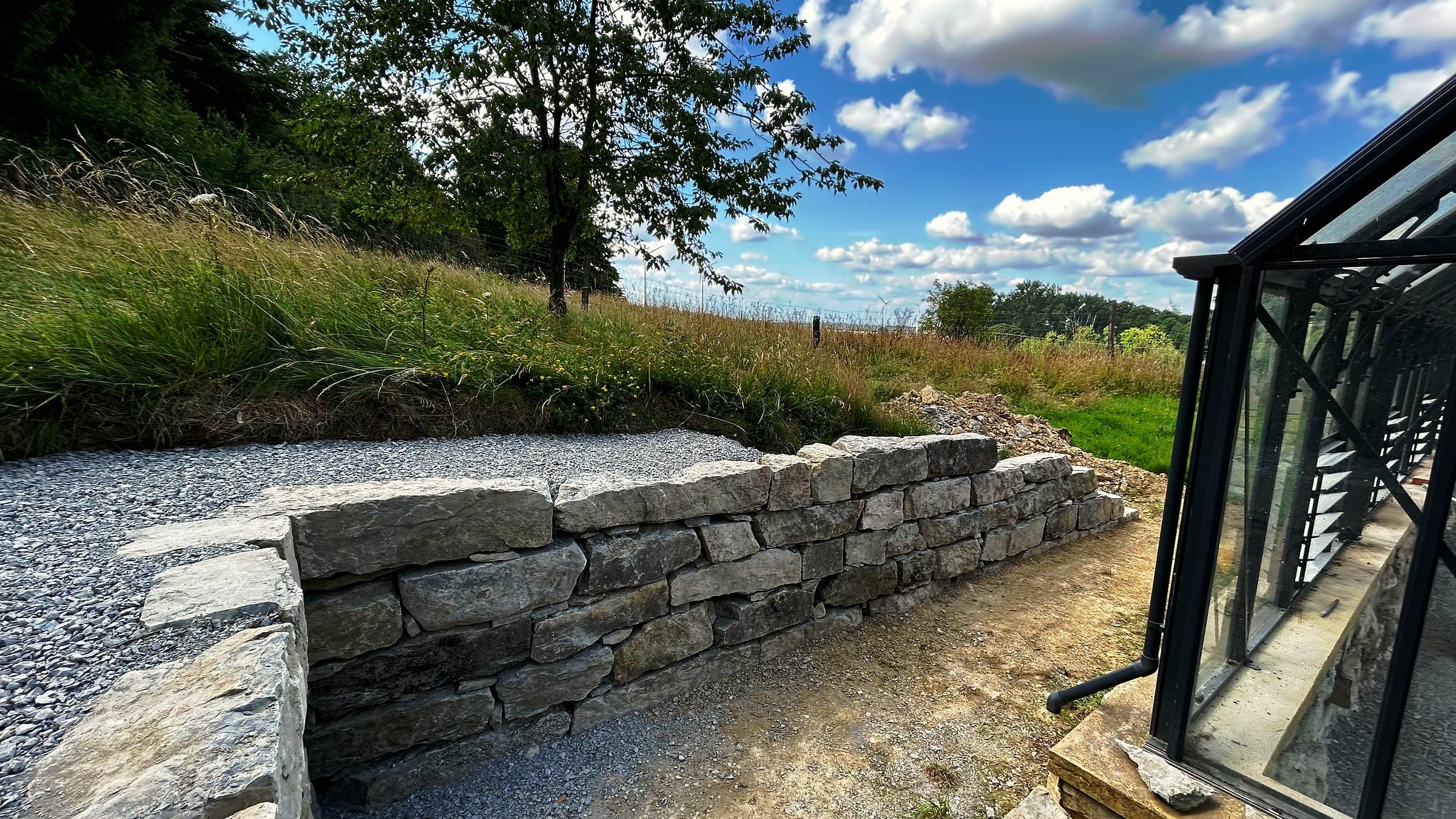 Neue Trockenmauer aus Naturstein am Hang neben einem Gewächshaus